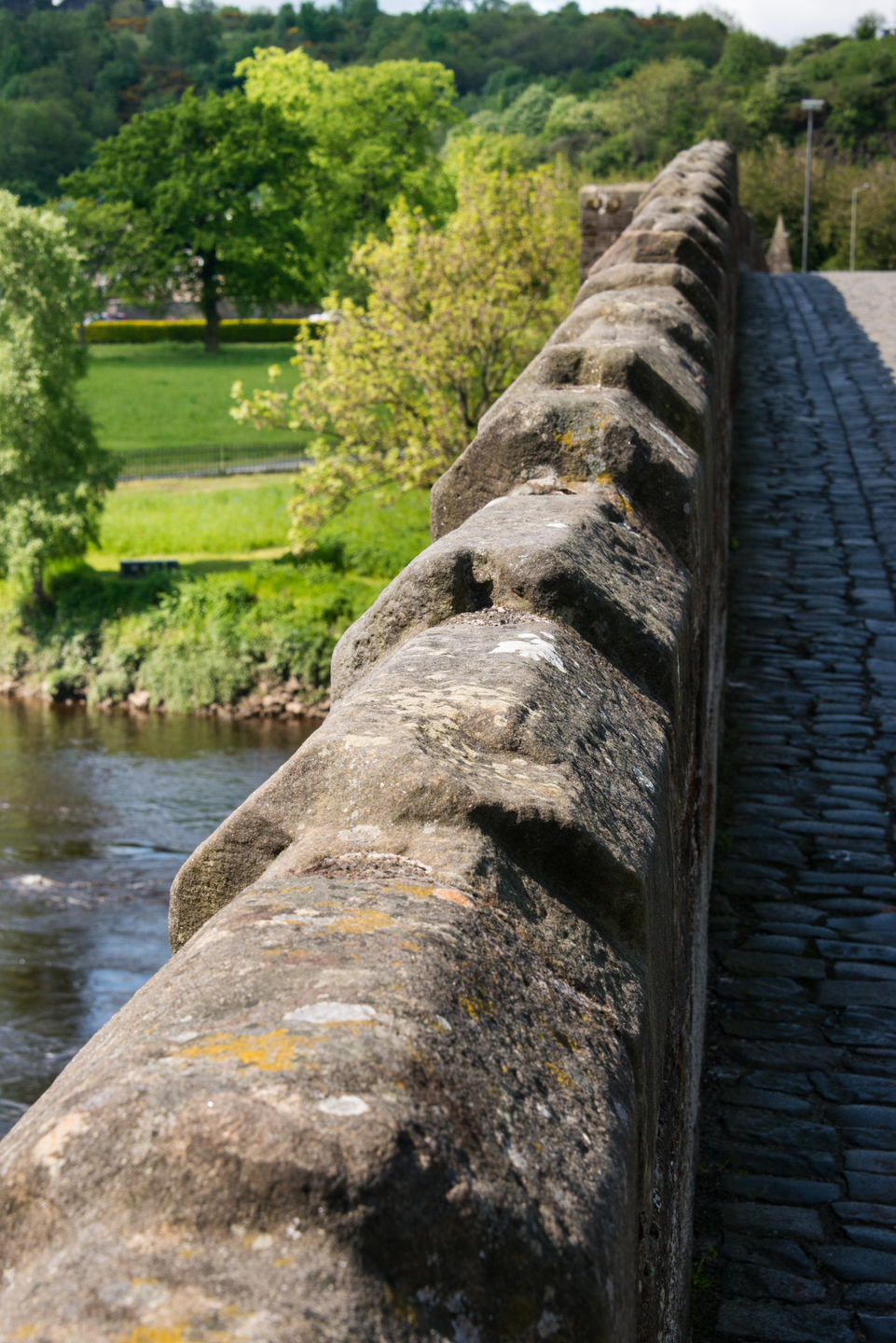 Close-up view along the stone parapet of Stirling Old Bridge, with the cobbled walkway on the right and the River Forth and green trees visible below