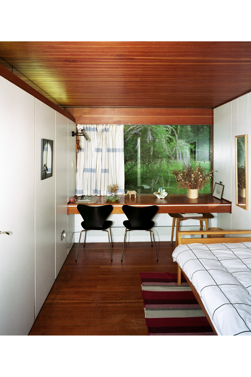 View of room with white panelled walls, wood panelled floor and ceiling, bed to the right, two chairs at a desk looking out to window with luscious greenery outside