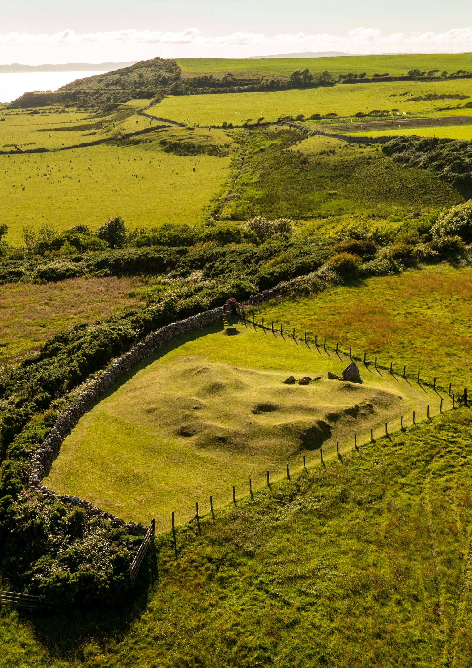 Aerial view of Torrylin Cairn, showing grass‑covered mounds and stone remains enclosed by a fence in the surrounding landscape.”