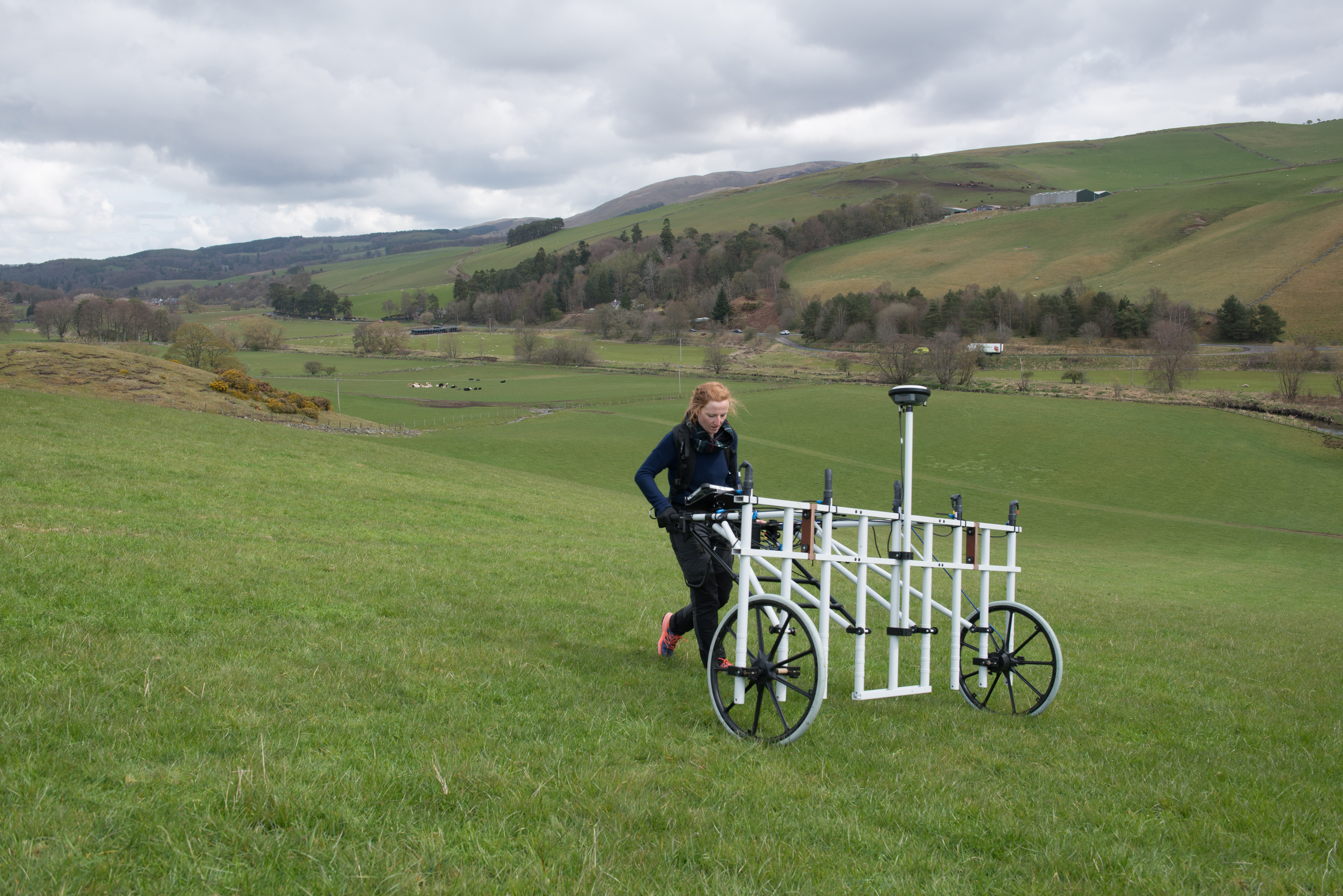 A woman pushing a geophysics device over grassland.