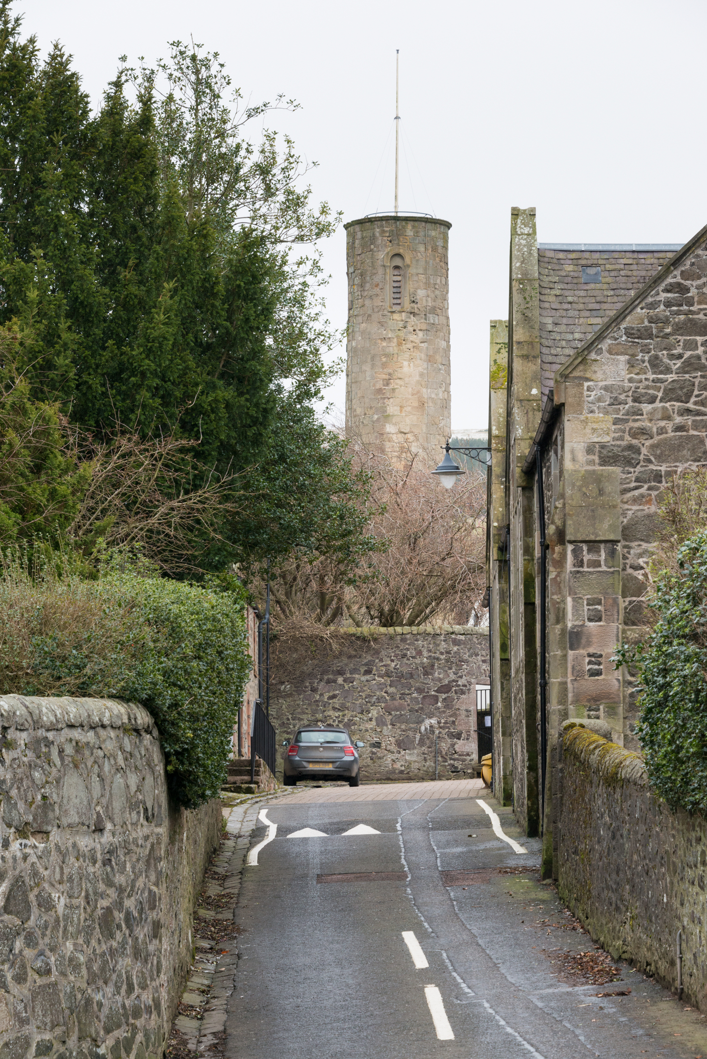 A distant view of Abernethy Round Tower rising above rooftops, seen from a narrow village lane lined with stone walls and houses.