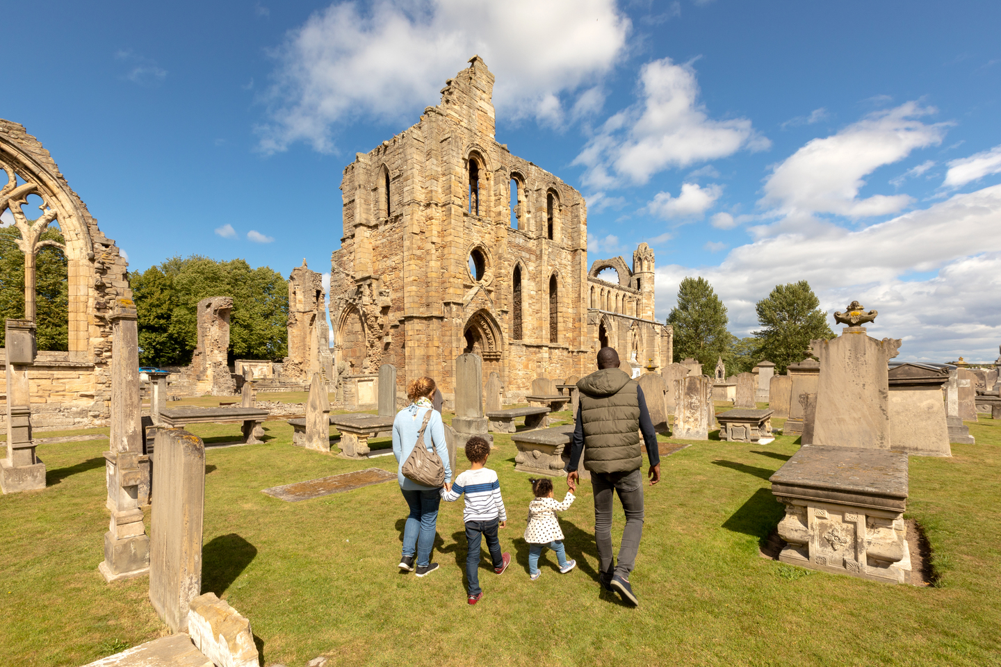 Two adults, holding hands with two young children, walk through a graveyard, with historical stone ruins in the background. It is a sunny day.