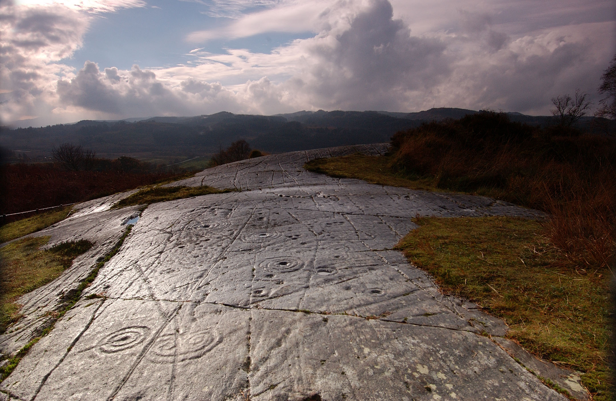 Wide stone slab with circular carvings and lines, grassy edge, hills and dramatic clouds in background.