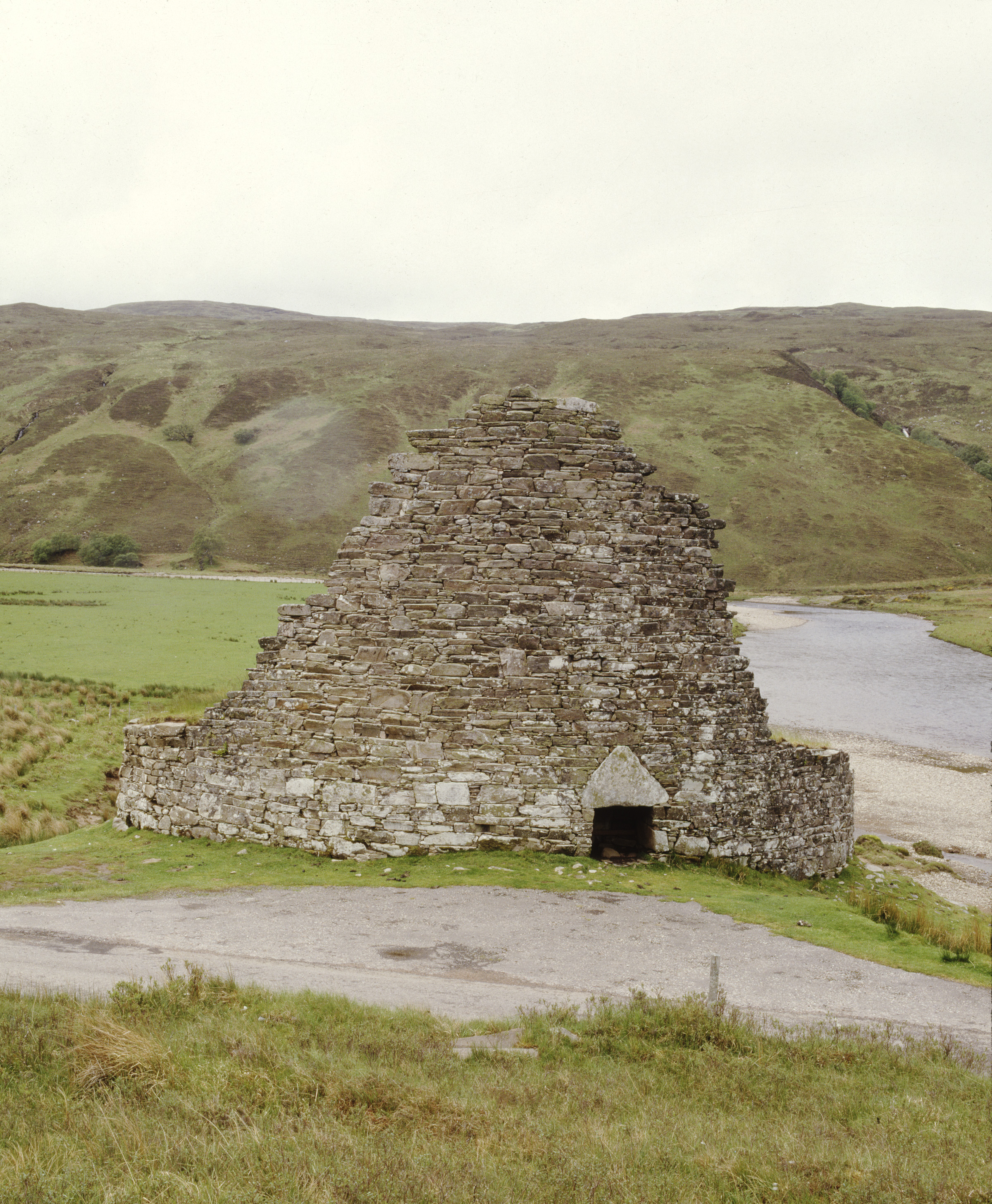 Front view of the ruined stone broch at Dun Dornaigil, showing its tall, triangular drystone wall and narrow entrance with a large triangular lintel stone. The structure sits on grassy ground beside a winding river, with rounded hills forming the backdrop under an overcast sky.