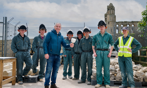 MSP Richard Lochhead stands with group of Historic Environment Scotland (HES) apprentices in a stonemasons' yard. He holds a mallet, while the apprentices wear personal protective equipment (PPE). The tower of Elgin Cathedral is in the skyline behind them.