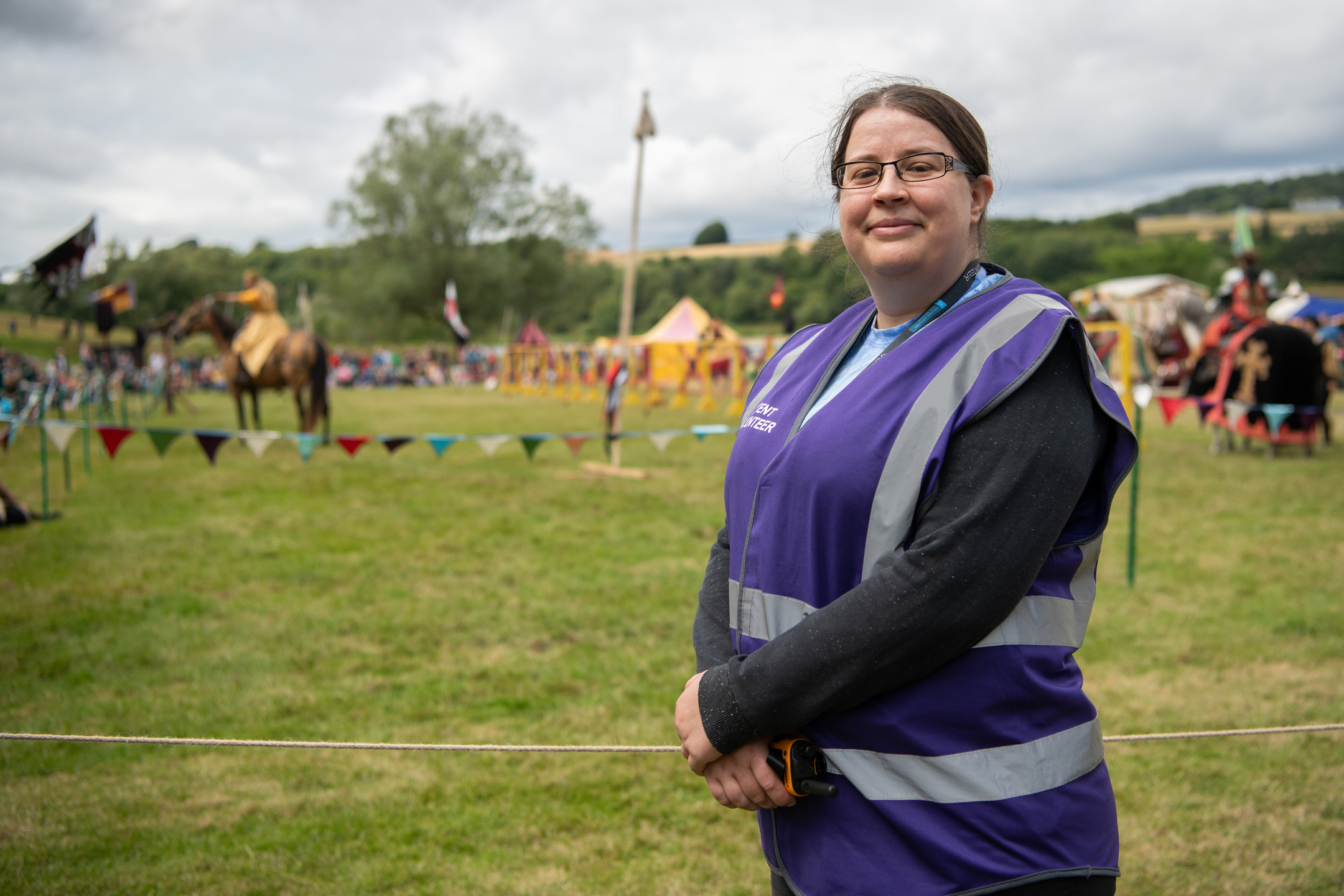 Person wearing a purple event vest stands in a grassy field in front of a roped-off arena, with a horse and colourful tents in the background.