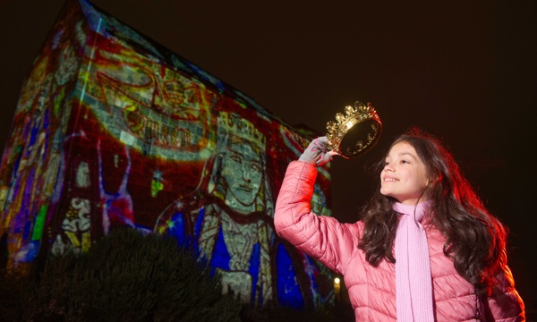 A young girl with long brown hair wearing a pink coat, pink scarf and grey gloves smiles whilst holding a gold crown over her head. Behind her is an old stone chapel with light projections shone onto its side. The projections show larger than life stained glass window depictions of a medieval queen. 