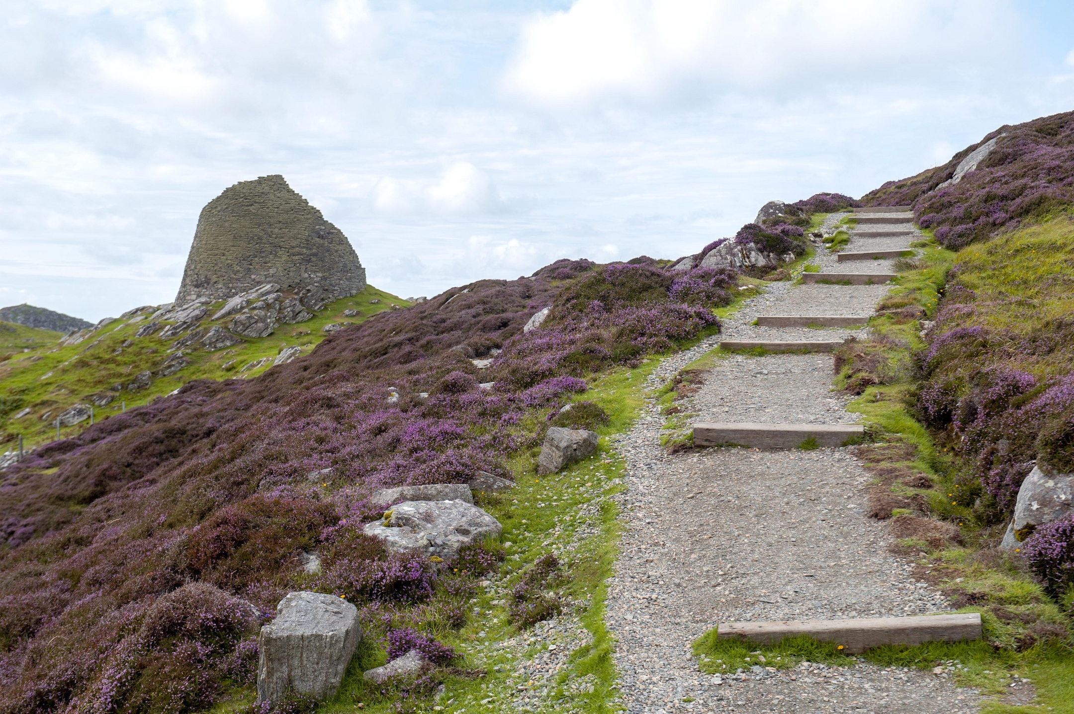 A well-maintained path weaves through a rocky hillside with a stone ruin on the left