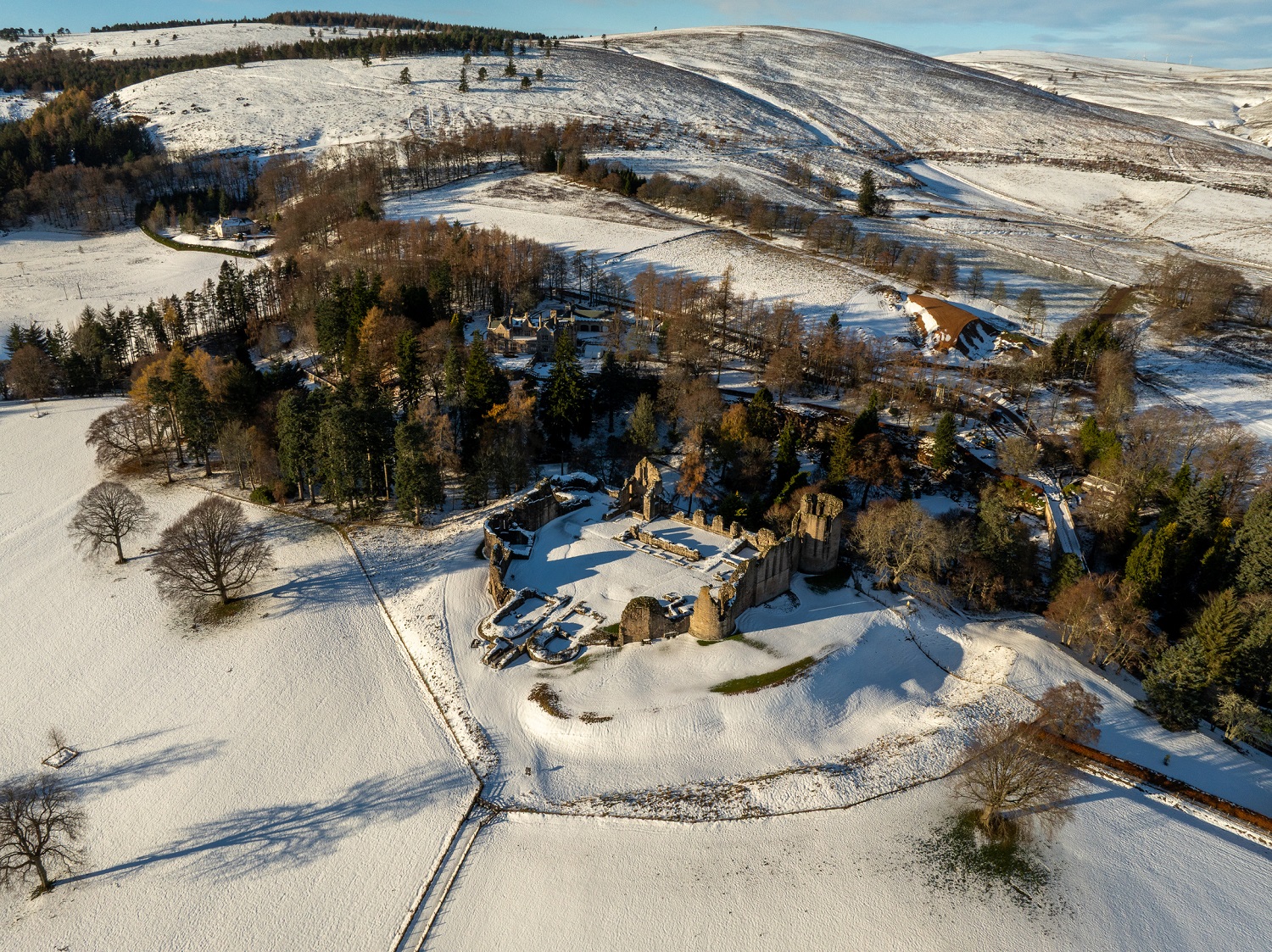 Aerial view of Kildrummy Castle in winter, showing the snow‑covered medieval ruins surrounded by open white fields and clusters of trees. Rolling hills in the background are also blanketed in snow under bright, clear daylight.