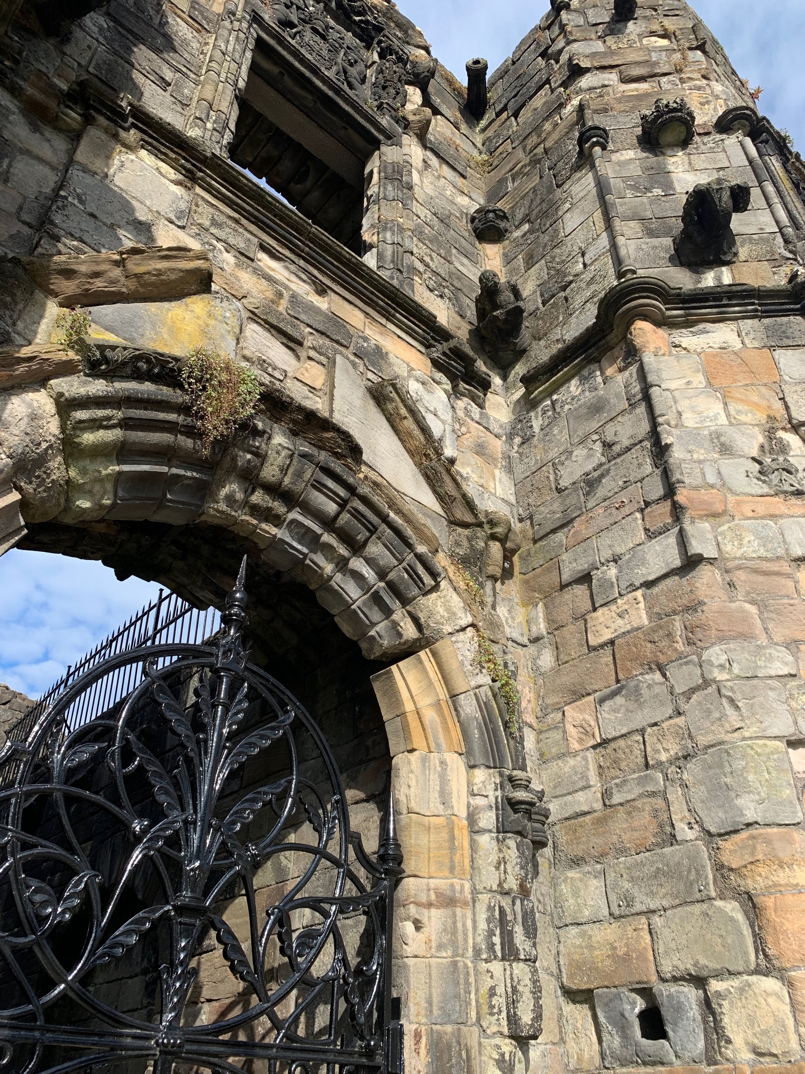 Close-up view of the main arched entrance of Mar’s Wark, a Renaissance-style ruin in Stirling, Scotland. The arch is constructed from weathered stone blocks with decorative moldings and carvings. Above the arch, remnants of ornate stonework and window openings are visible. A black wrought-iron gate with intricate scrollwork fills the archway, and patches of moss grow between the stones.