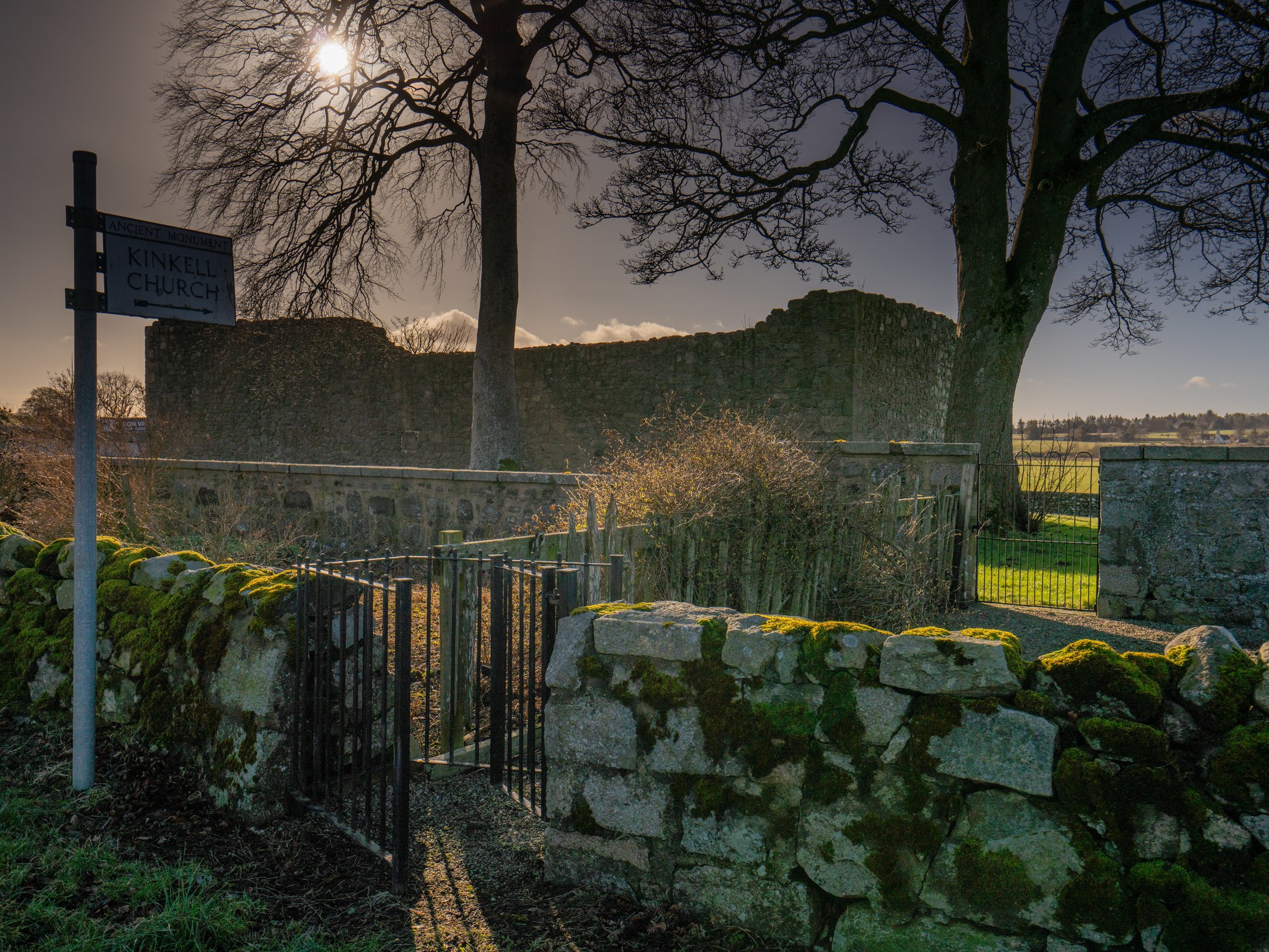Entrance gate and moss-covered stone wall leading to the ruins of Kinkell Church at sunset.