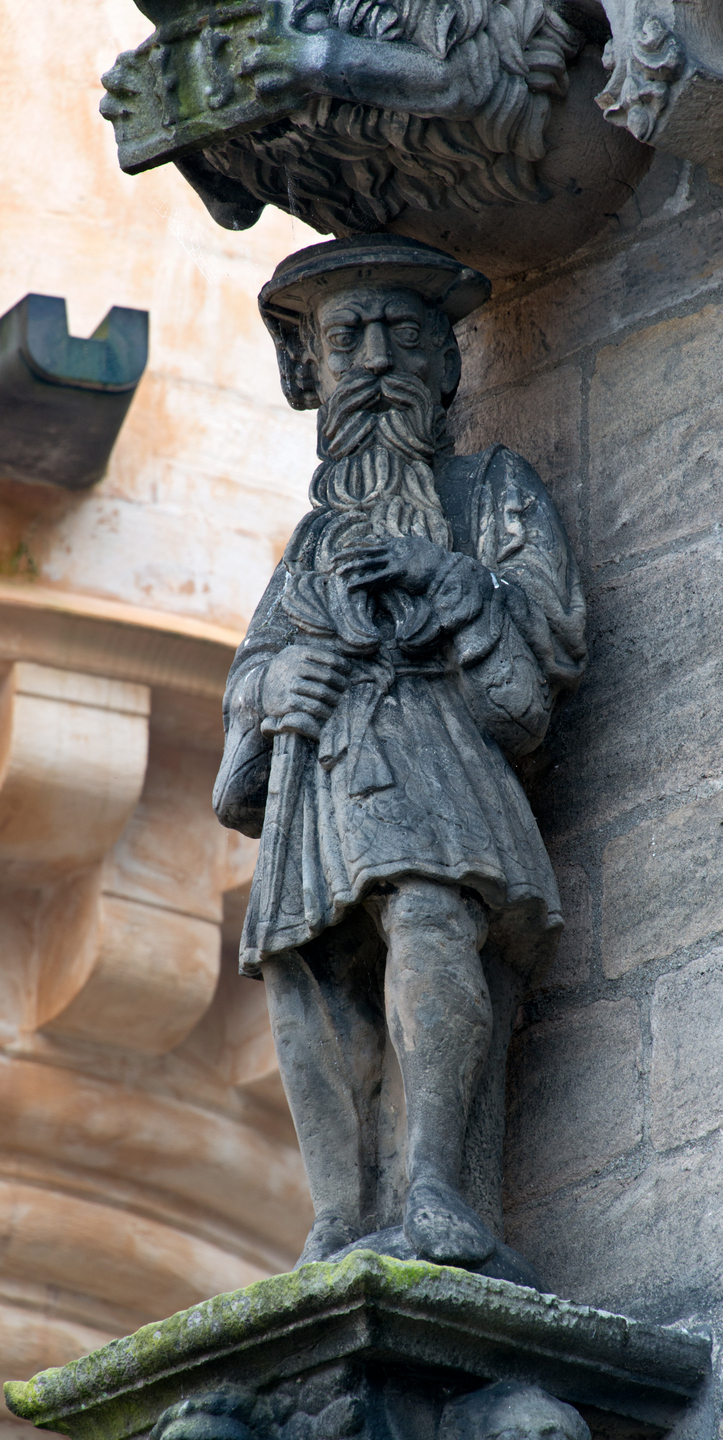 A statue of a man sits on a platform, which is connected to a wall. He has a hat on, a long, curly beard and wears a royal tabart.