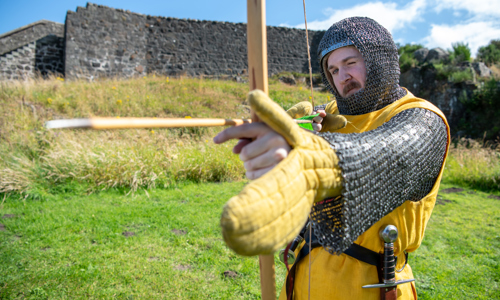 A man with a moustache dressed in medieval costume holds a bow and arrow. He faces the camera and has one eye closed as he aims to arrow. Behind him is green grass and an old stone wall, with a blue sky above and the sun shining.