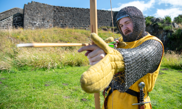 A man with a moustache dressed in medieval costume holds a bow and arrow. He faces the camera and has one eye closed as he aims to arrow. Behind him is green grass and an old stone wall, with a blue sky above and the sun shining.