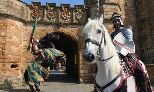 A knight dressed in armour is holding a lance and riding a white horse in the foreground of the photo. In the back there is a knight holding a green and gold flag, riding a black horse which is dressed in green, gold and red horse blanket. The horse is rearing up with his front hooves off the ground. The knights are in front of the archway entrance to Linlithgow Palace, which features four colourful coats of arms on the stonework.