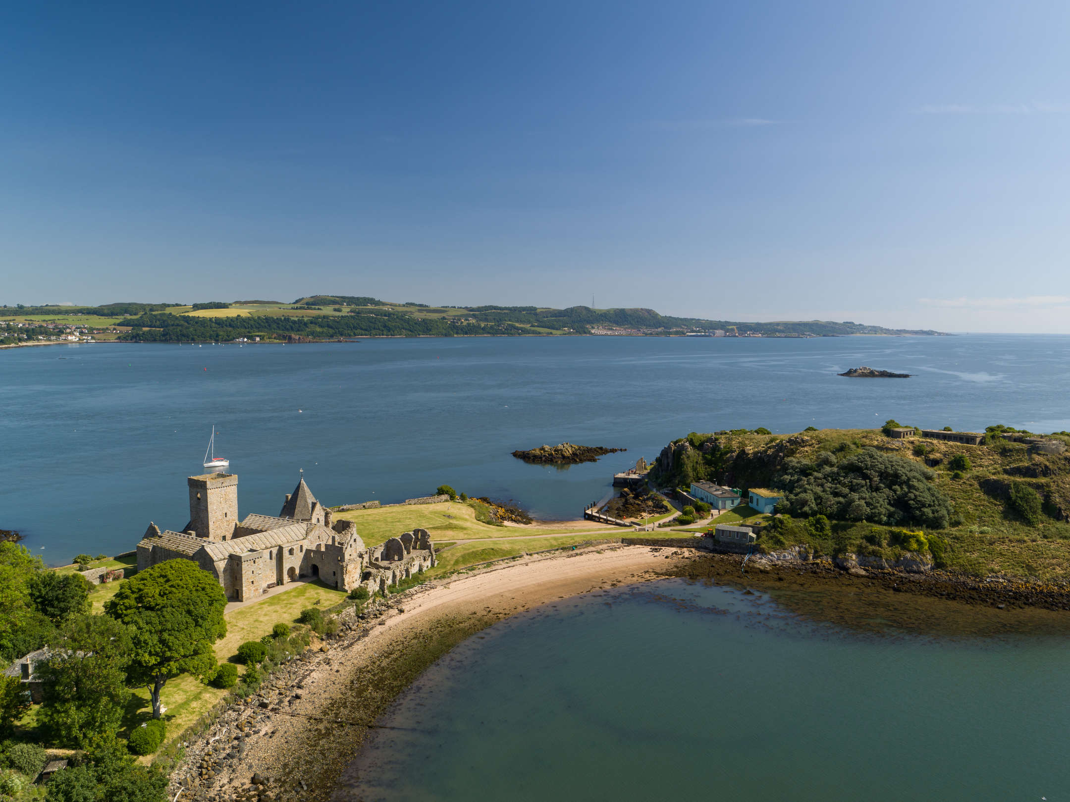An aerial view of a large stone historic abbey on a small grassy island with a sandy beach, trees and a boat in the water nearby