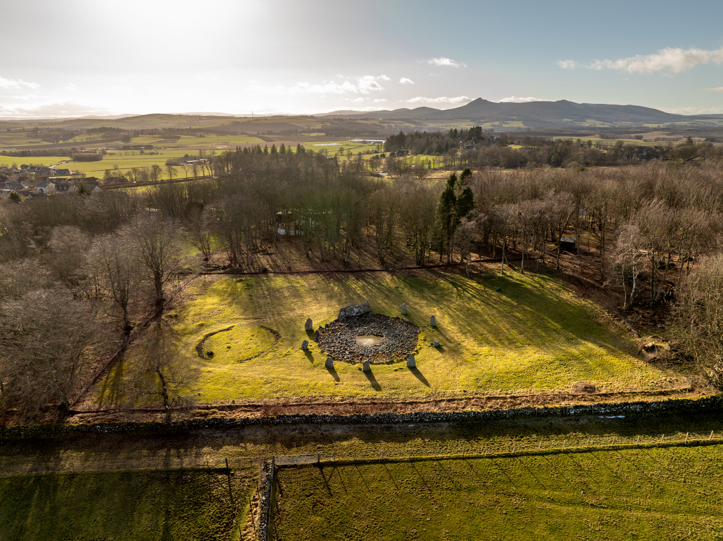 A field with trees surrounding it, in the centre there is a circle filled with stones, with larger stones surrounding it.