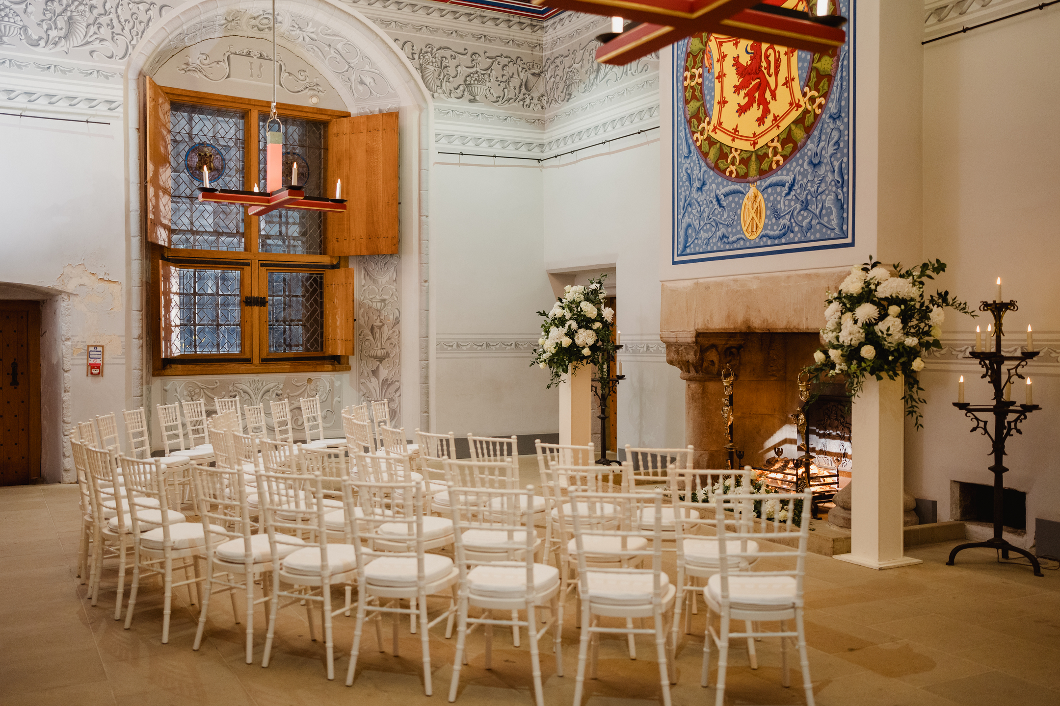 Rows of white chairs arranged for a ceremony in a historic room featuring a heraldic painting, stone fireplace, arched windows and floral arrangements. 