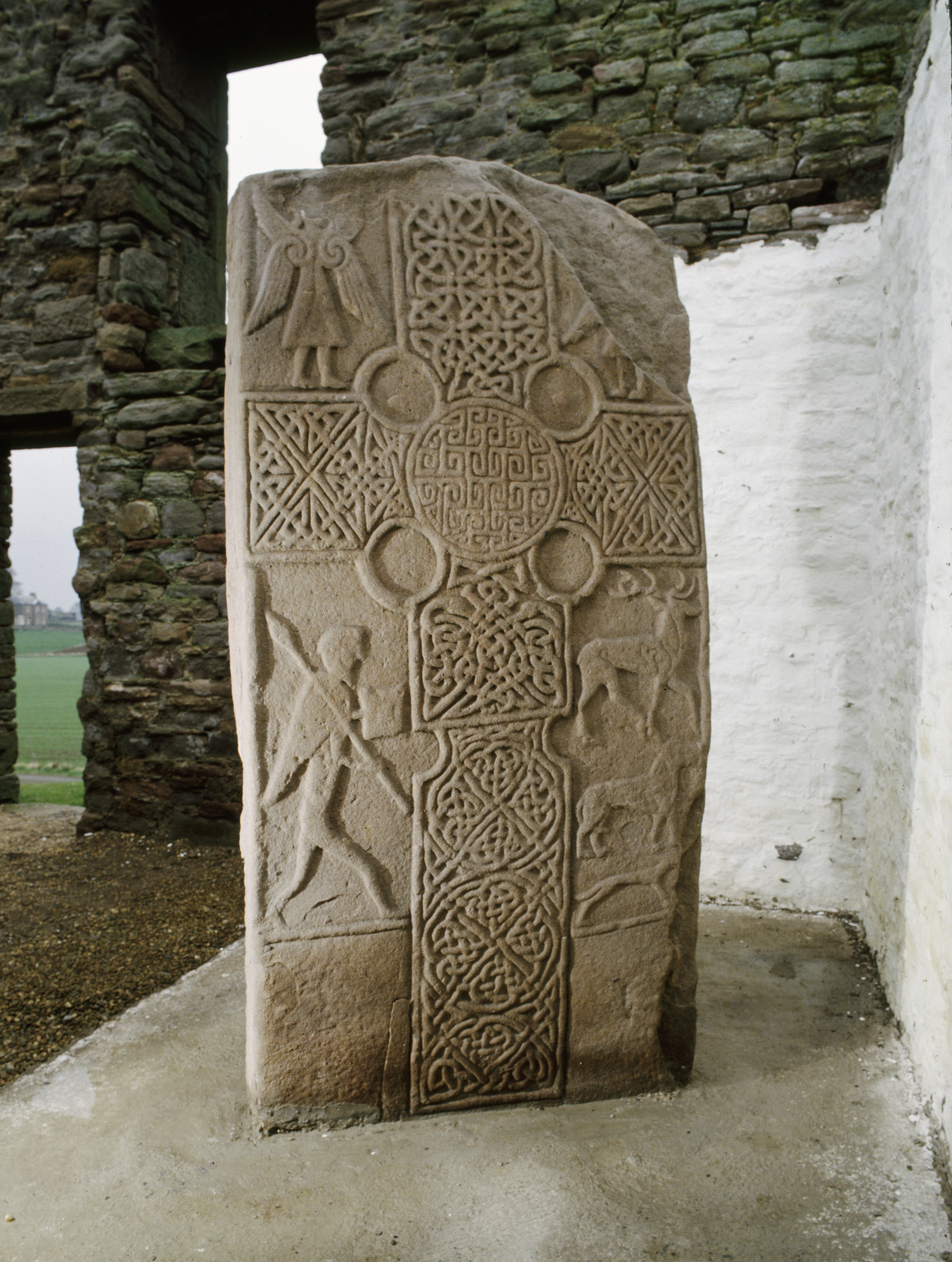 Full view of the Eassie Pictish sculptured stone standing upright. The tall sandstone slab is carved with an ornate cross, featuring knotwork panels, circular bosses, and interlaced patterns. Below and beside the cross are carved human and animal figures, including a standing figure with a staff and another leading an animal. The surface of the stone is weathered but well‑defined, lit to highlight the detailed carvings. The background shows rough stone walls with open window spaces.