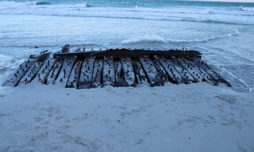 Old wooden timbers with rusty bolts partially buried in sand at the edge of a calm ocean shore with gentle waves.