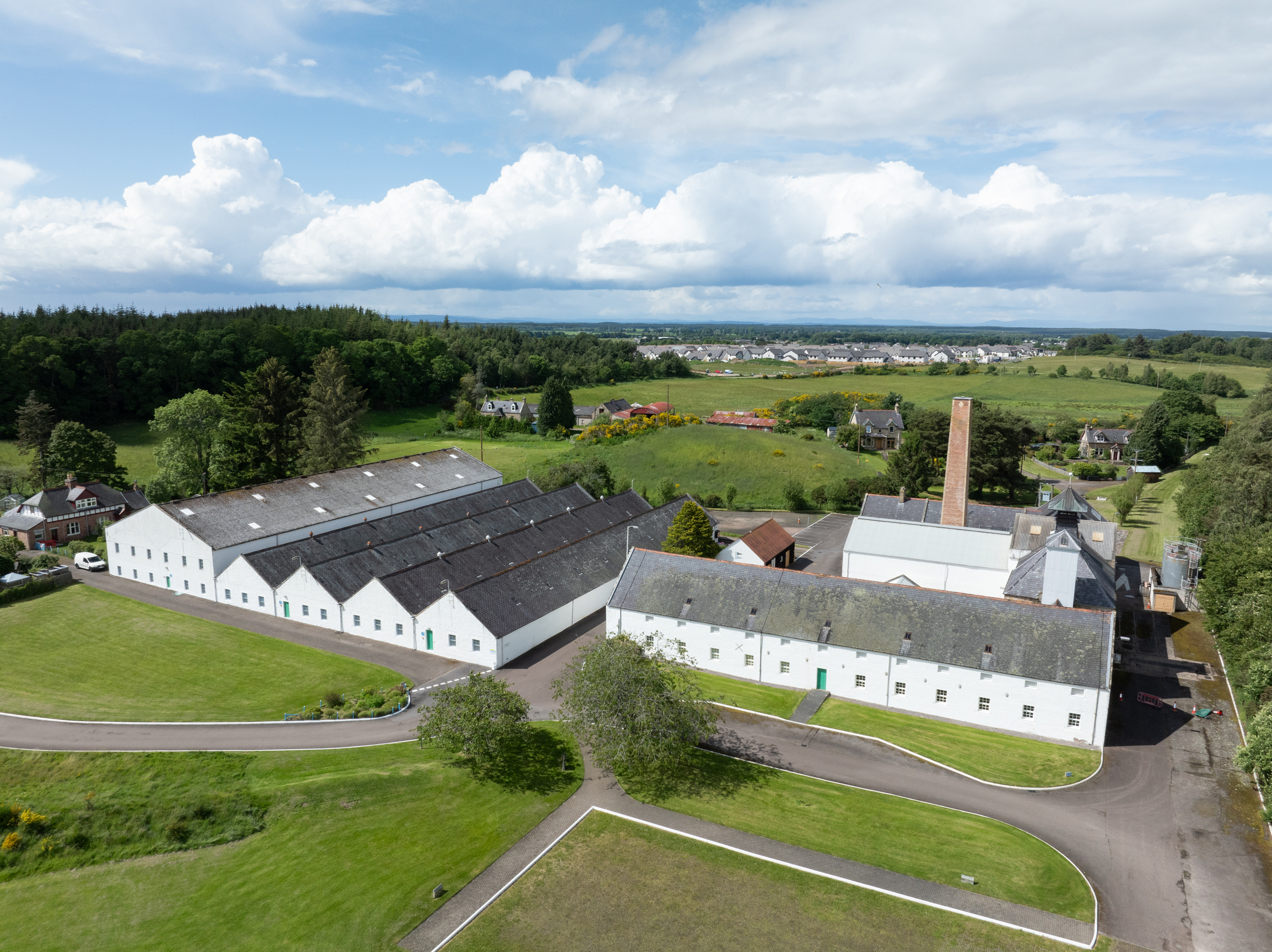 Aerial view of Dallas Dhu distillery showing white warehouse buildings, landscaped grounds and surrounding countryside under a bright summer sky.