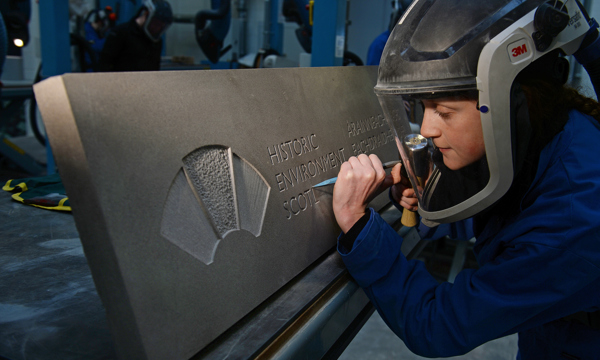 A stonemason carves the Historic Environment Scotland logo into stone.