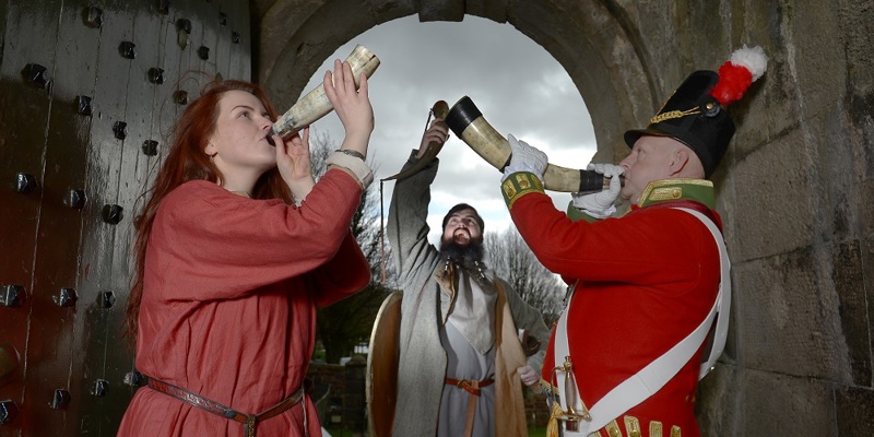 Three re-enactors blowing horns under an archway