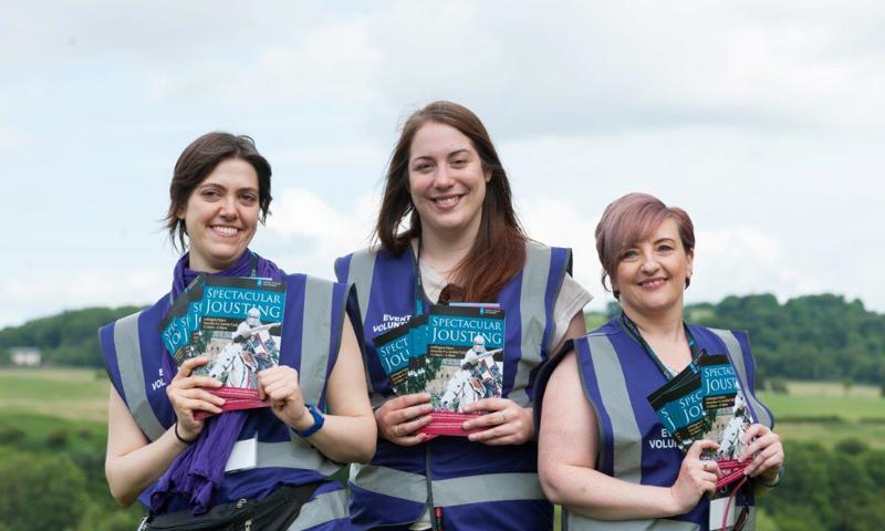 Three volunteers with Spectacular Jousting programmes, wearing purple vests