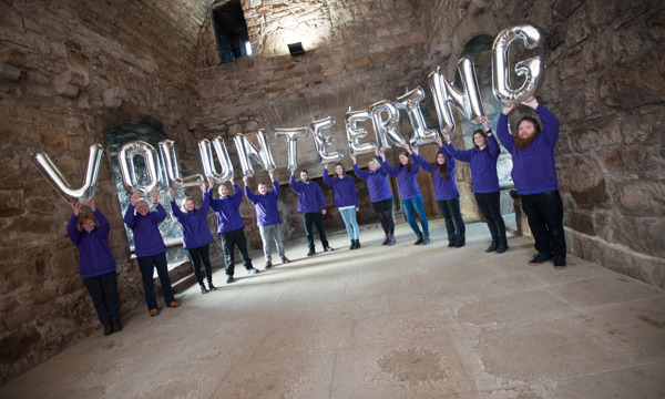 Twelve volunteers in purple fleeces, each holding up above their heads a silver coloured inflatable letter, together spelling the word VOLUNTEERING