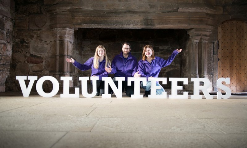 Three volunteers in front of a fireplace, behind large lettering spelling the word VOLUNTEERS