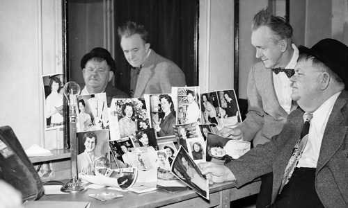 Laurel and Hardy, one seated, in a room, looking at photos arranged in front of a large mirror