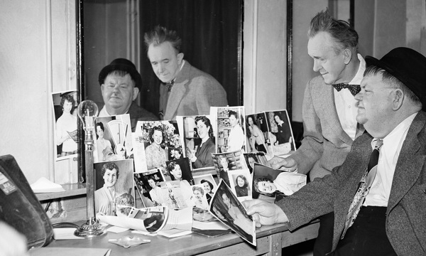 Laurel and Hardy, one seated, in a room, looking at photos arranged in front of a large mirror