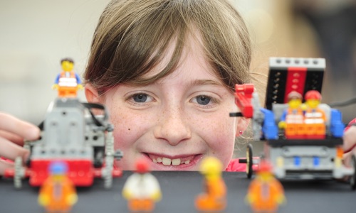A close up of a girl's face as she crouches behind a table, playing with Lego models