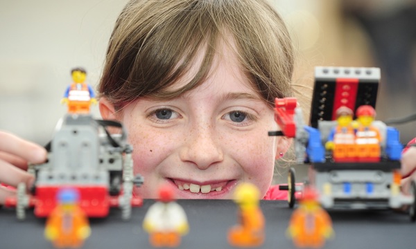 A close up of a girl's face as she crouches behind a table, playing with Lego models