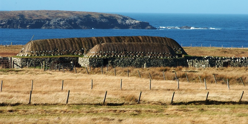 Distant photo of The Blackhouse at Arnol with fields in the foreground and the North Atlantic Ocean in the background