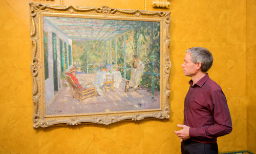 A photograph of a man in a red shirt standing beside a painting of people sitting and standing in a pergola