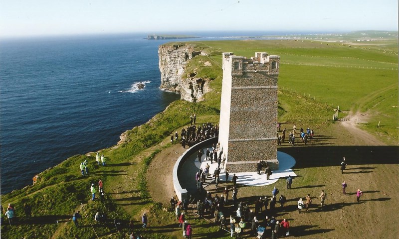Aerial view of the Kitchener Memorial in Orkney, surrounded by visitors, with the cliffs and the sea visible at the left hand side