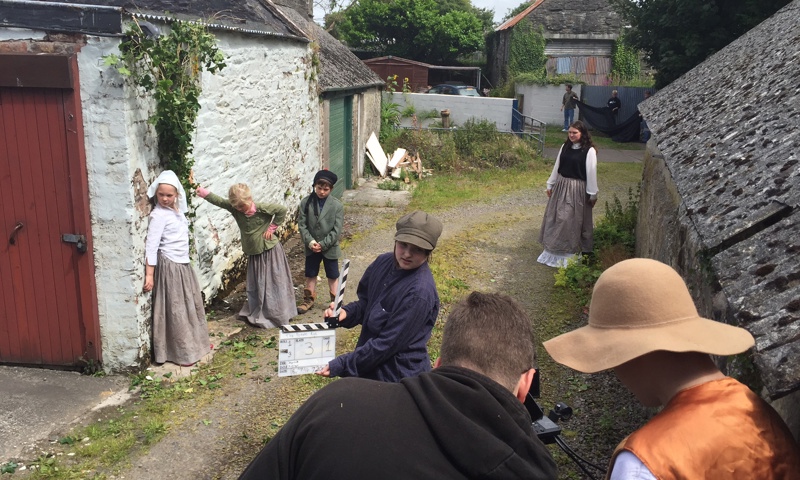 Filming of children in traditional clothing hiding behind the wall of a shed