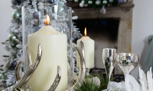Large candles in glass containers with silver antler decorations, next to silver glasses, with a fireplace in the background