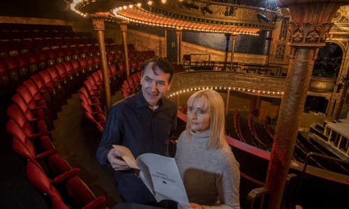 A man and a woman in the upper balcony of a theatre, holding a document titled "What's Your Heritage?"