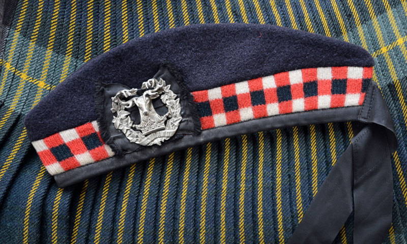 A photograph of a navy blue wool cap with a red and white check band and a badge in the centre