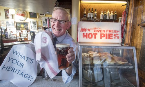 Man behind a counter holding a pint of beer, next to a cabinet of hot pies, with a sign that says "What's your heritage?"