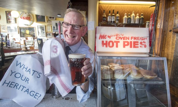Man behind a counter holding a pint of beer, next to a cabinet of hot pies, with a sign that says "What's your heritage?"