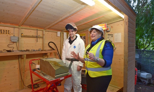 A photograph of a woman in a high visability jacket and hard hat, standing with a man in white overalls beside large stone block.