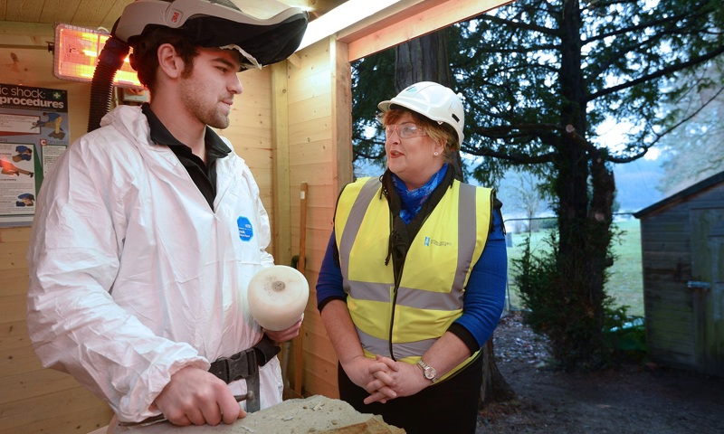 A photograph of a young man in white overalls sood beside a woman in a high visibility jacket and a hard hat.