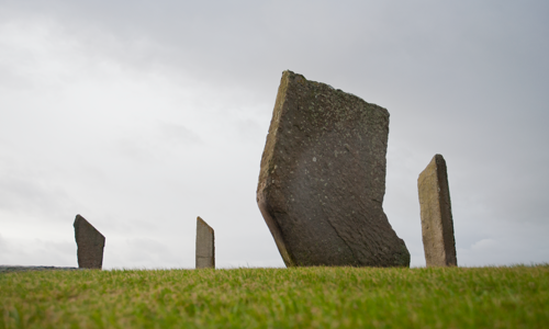 A photograph of standing stones on green grass with a cloudy sky behind.