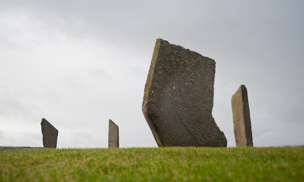 A photograph of standing stones on green grass with a cloudy sky behind.