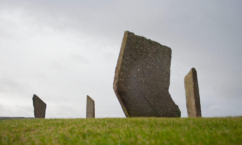 A photograph of standing stones on green grass with a cloudy sky behind.