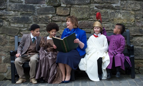 A woman on a bench reading to 4 children wearing re-enactment clothing at Edinburgh Castle