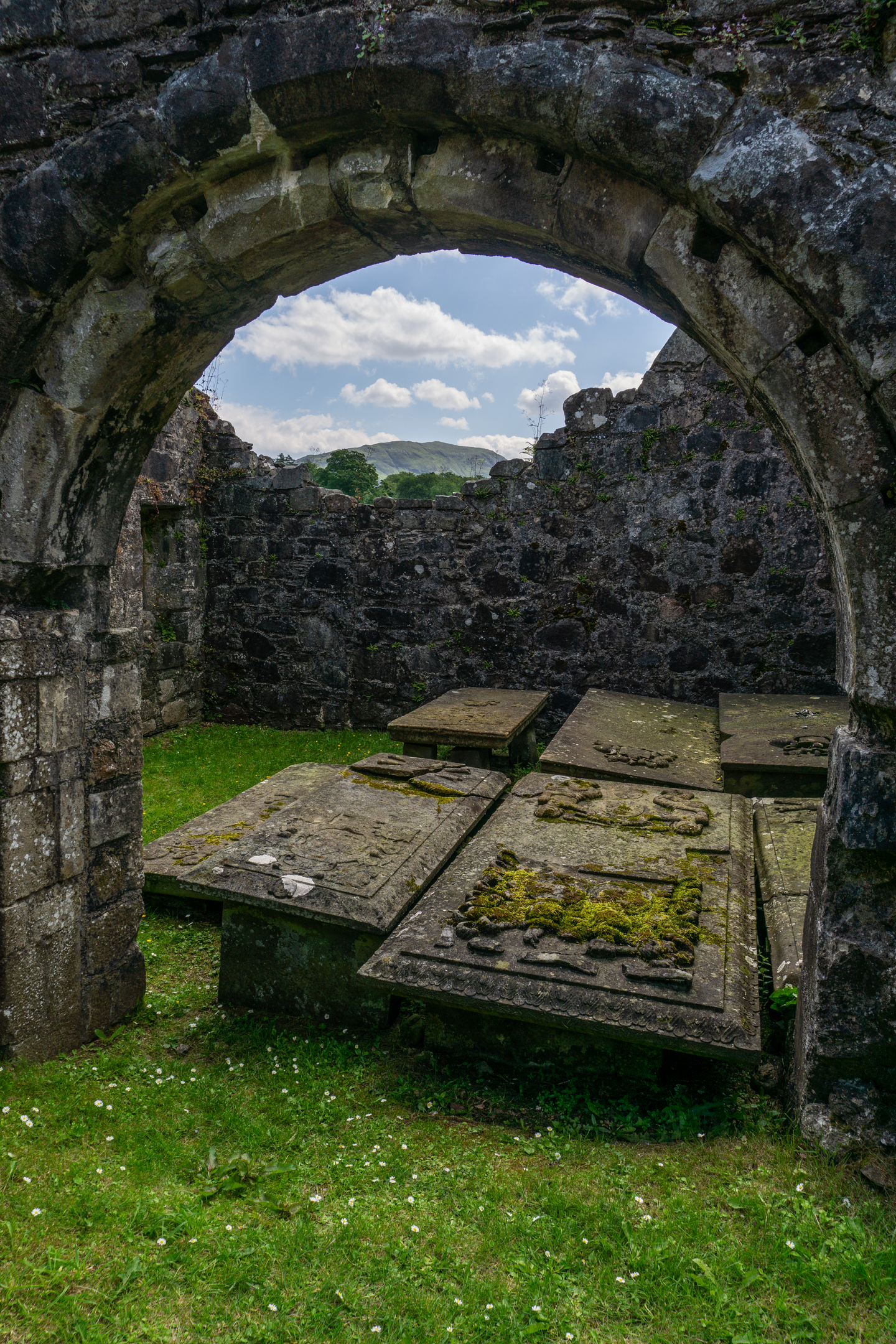 A view through a rounded stone archway into a small enclosed space containing several flat medieval tomb slabs covered in moss, with a glimpse of hills and sky through a gap in the far wall.